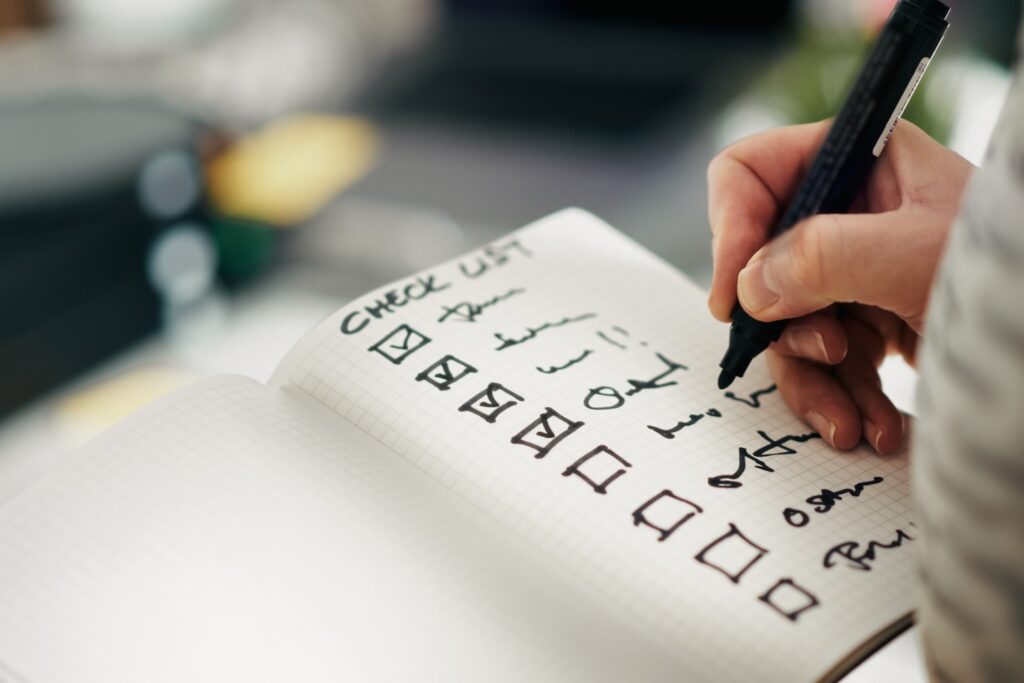Checklist notebook task progress accuracy. Close-up hand holding pen while marking completed tasks grid notebook. Symbol of productivity, precision, quality control, and workflow management in business or personal planning